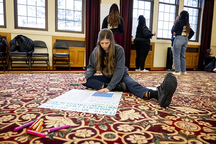 A female college student sits on the floor while writing on a large piece of paper.