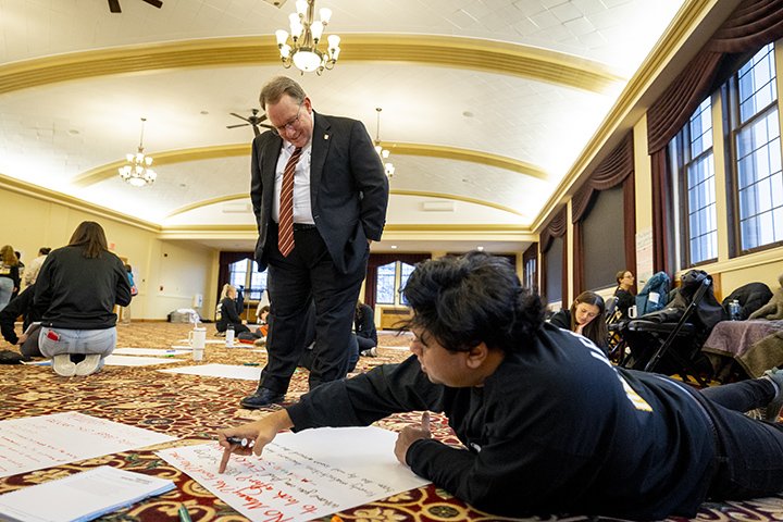 A male student lies on the floor next to a large piece of paper while explaining what he wrote as CMU President Bob Davies looks on.