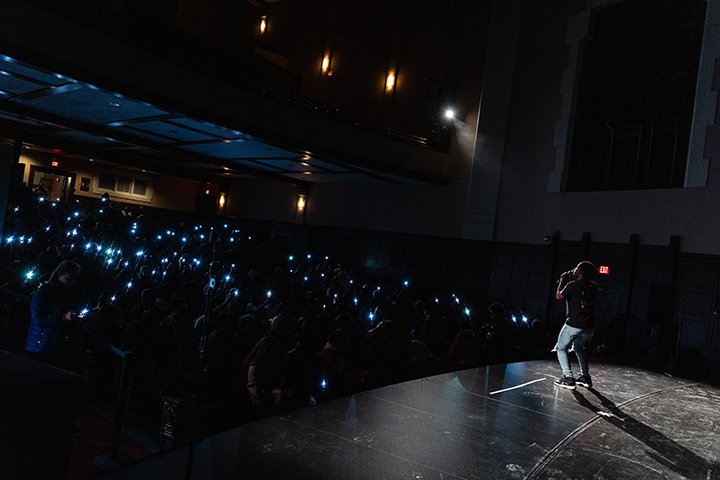 A person stands on the stage at Plachta Auditorium while the dimly-lit crowd holds up their mobile phones while waving their hands in the air.