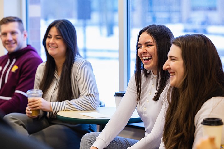 Four students sit at a small round table while smiling and drinking coffee.