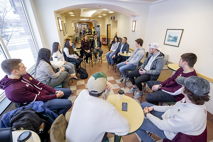 A wide-angle shot of nearly a dozen students sitting in a circle inside a coffee shop.