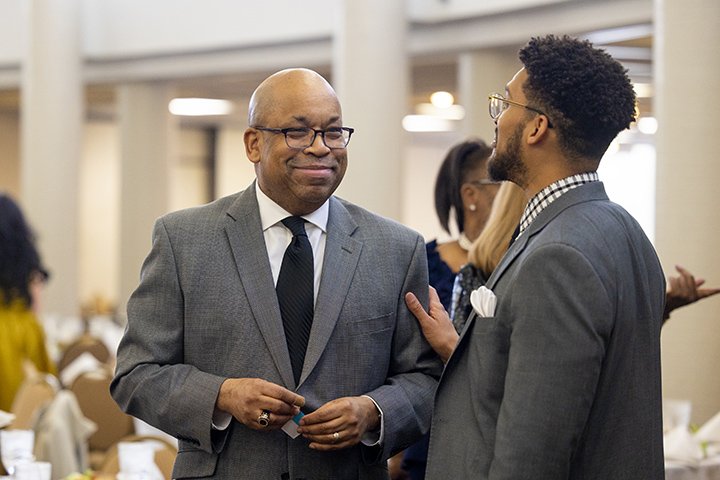 Two men wearing suits talk while standing in the University Center's Rodunta Room.