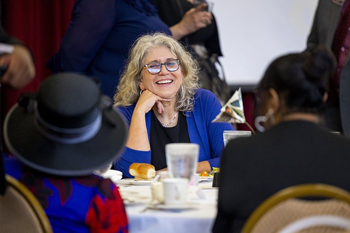 A woman wearing a blue blazer and glasses sits at a table smiling while talking to others.