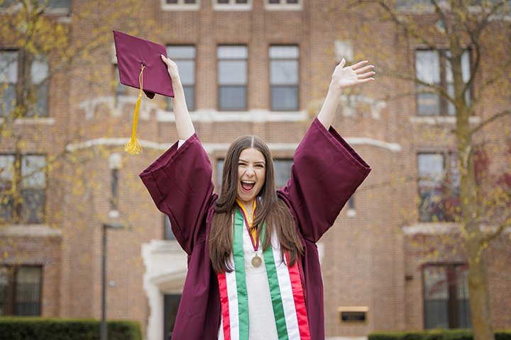 A female college student wearing her commencement attire raise her hands in the air while holding a graduation cap and smile mouth wide open.