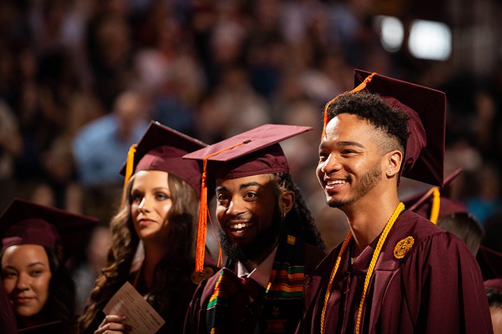 Three college graduates stand together during commencement looking at the stage.