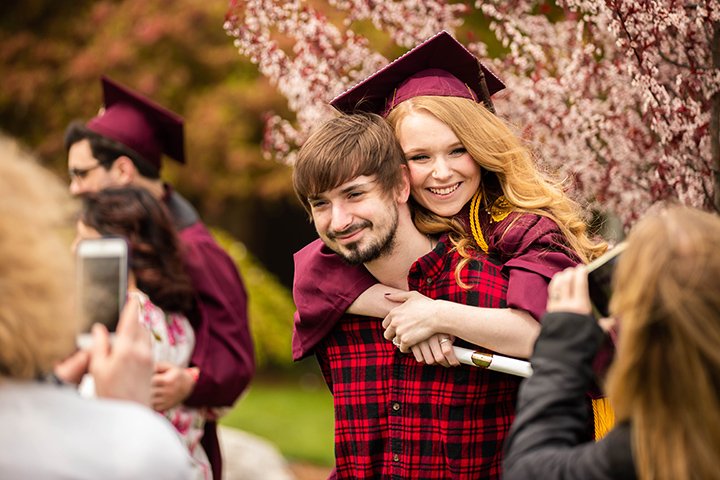 A female college student holds onto a young man's back, smiling at the camera.