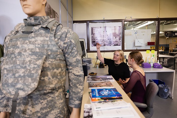 A professor and a student sit at a table discussing fashion design as a mannequin in an army uniform stands in the foreground.