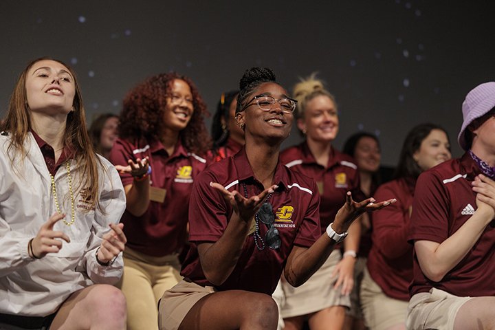 A group of college students wearing CMU attire stands on a stage singing as part of new student orientation.