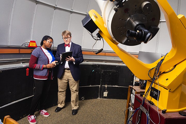 A student and a faculty member stand near a large telescope inside the Brooks Astronomical Observatory.