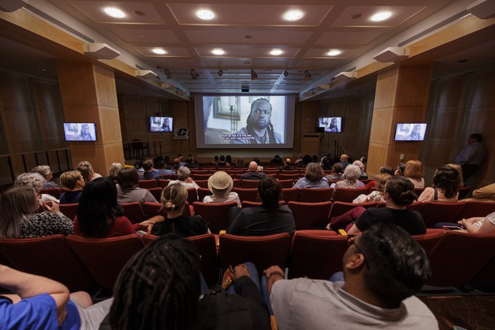 A large group of people sit in an auditorium looking at a movie screen showing a documentary as four televisions flank the sides of the room showing the same documentary.