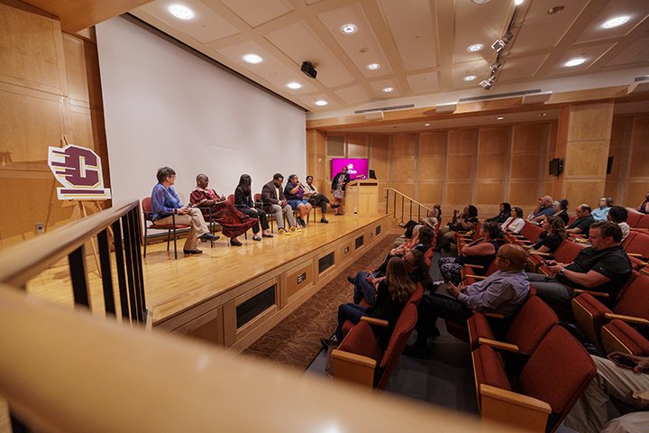 Six people sit in chairs on the stage of an auditorium with a host standing near a podium off to the side. A large group of people looks on from their seats.