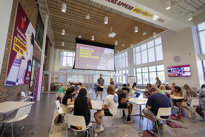 Approximately 25 soon-to-be college freshmen sit in groups of three at small, square tables for College of Business Administration orientation inside the brightly-lit atrium of Grawn Hall.