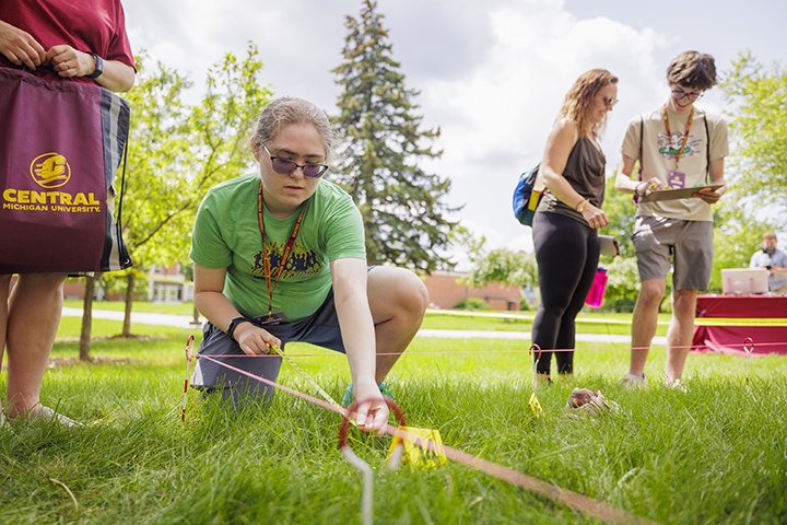 A high school student in a green shirt and sunglasses crouches in the grass at a mock crime scene holding out a tape measure.