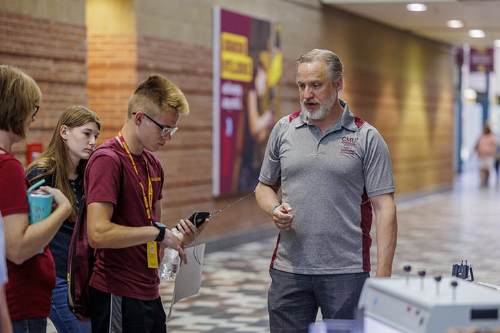 A CMU faculty member in a gray polo shirt talks to three high school students in a well-lit atrium area.