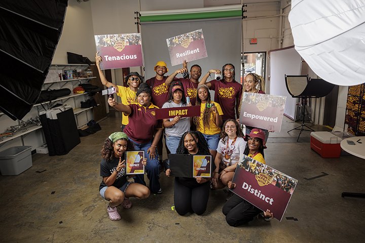 12 CMU students wearing CMU clothing pose for a group photo inside a large room. Some students hold signs with inspiring words on them, while others hold cameras.
