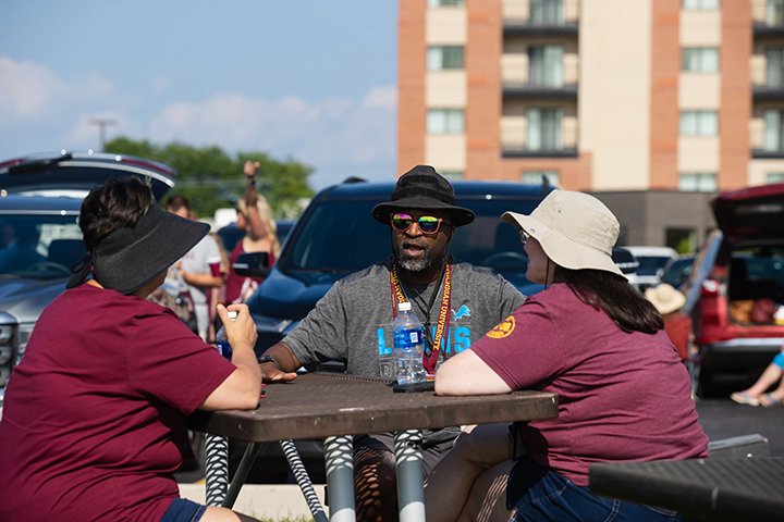 Three people wearing hats sit at a picnic table talking during a tailgating event.