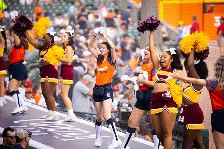 Cheerleaders dressed in maroon and gold CMU outfits and orange and navy blue Detroit Tigers colors stand on top of a baseball dugout and engage the crowd.