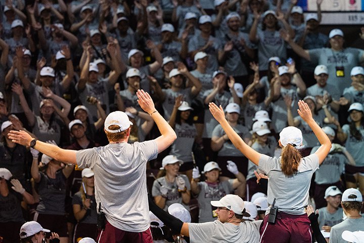 Two students in white hats, gray shirts and maroon shorts have their backs to the camera and their arms raised as they stand on stage urging a large crowd to wave their arms in the air.