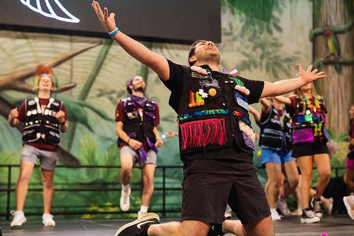A student in black shorts, black shoes and a black safari vest is on his knees on stage with his arms wide open as a group of similarly dressed students sings behind him.