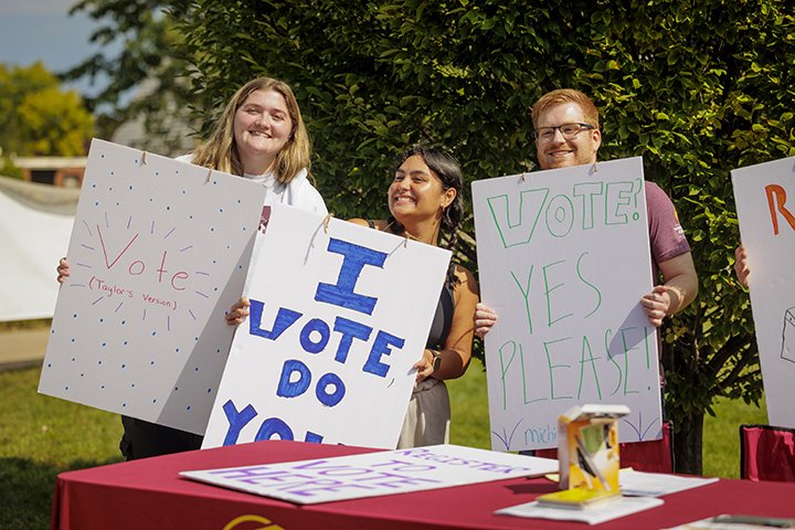 Three students stand behind a table holding large signs encouraging people to register to vote.