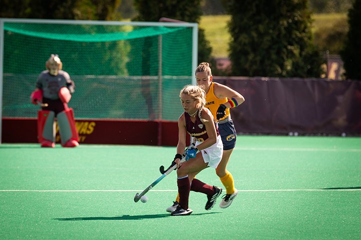 A CMU women's hockey player controls the ball on the field as a defender closes in on her to make a play. In the distance, a goal keeper stands ready.