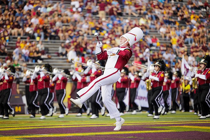 CMU's drum major high steps on the football field with wearing a large white hat as the band plays in the background.