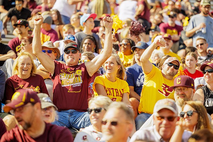 A closeup of a group of four football fans in the stands at a CMU football game, two men wearing hats and sunglasses pump their arms in the air while the women on either side of them laugh.