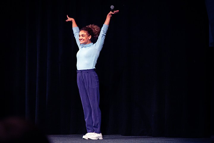 Former US Olympic gymnast Laurie Hernandez stands in front black curtains on a stage while striking a gymnastics victory pose.