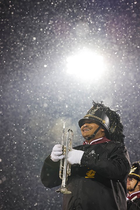 A CMU marching band trumpet player smiles and stands at attention as snow falls past the bright stadium lights.