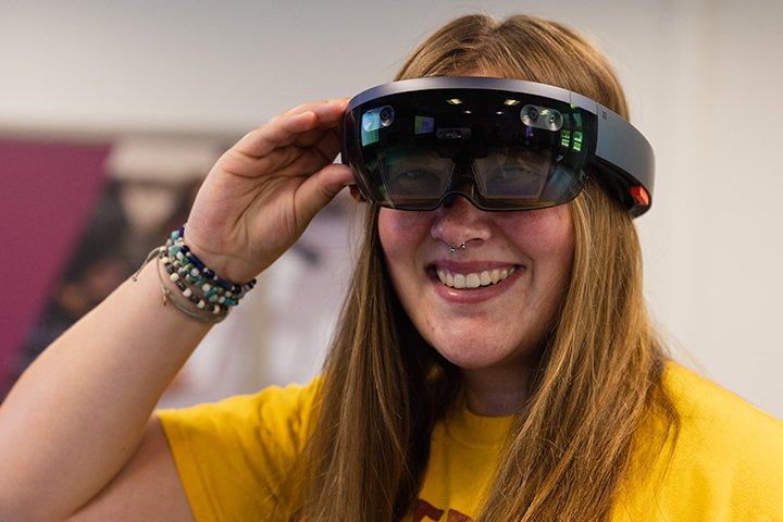 A female student wearing a gold-colored shirt and beaded bracelets smiles while wearing a black augmented reality headset.