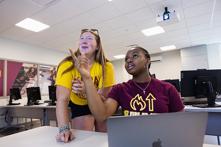 Two female students, one in a gold CMU shirt and the other in a maroon CMU shirt, look towards the screen in a classroom as they work on a laptop in a classroom.