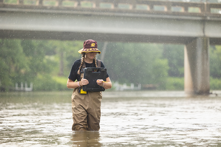 As rain falls, a young woman wades through a river wearing a floppy CMU hat and searching for mussels.
