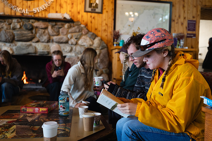 Biology students sit in chairs inside a lodge as a fire burns in the fireplace in the background.