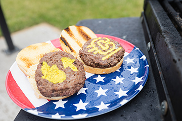 An American flag themed plate has two hamburgers on buns sitting on it. On one patty is painted the fire up emojis. On the other, the CMU action C.