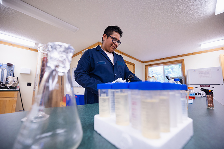 A student wears a dark blue lab coat in a lab as a beaker and test tubs sit on a table in the foreground.