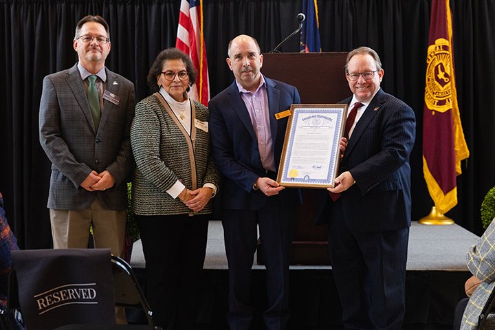 A man in a suit and a woman in professional attire stand next to two men in suit jackets holding a framed proclamation in front of an American flag.