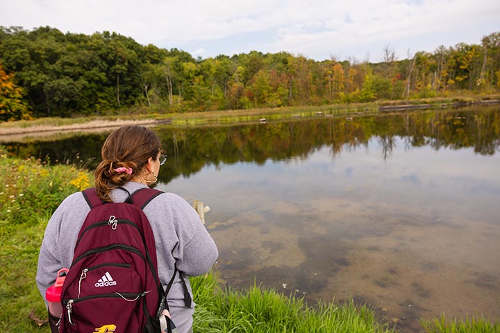 A woman with brown hair pulled back into a bun wearing a gray sweatshirt and maroon backpack looks off into a pond.