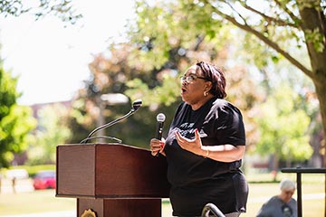 A woman in glasses wearing a black T-shirt speaks into a microphone while standing next to a podium on a sunny day.