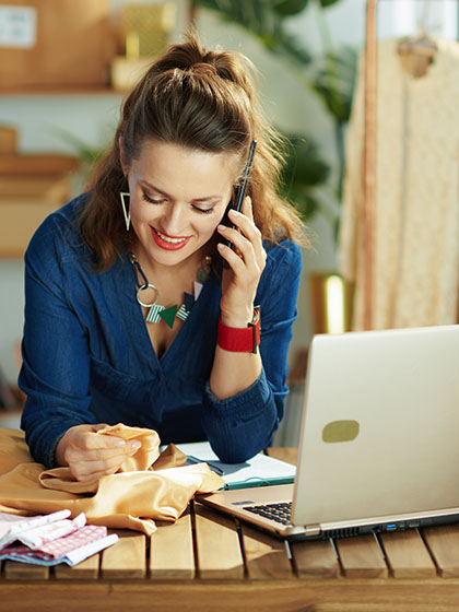 A woman in a blue blouse with brown hair partially pulled back smiles while talking on a phone and leaning against a counter containing folded fabric, boxes, and a laptop.