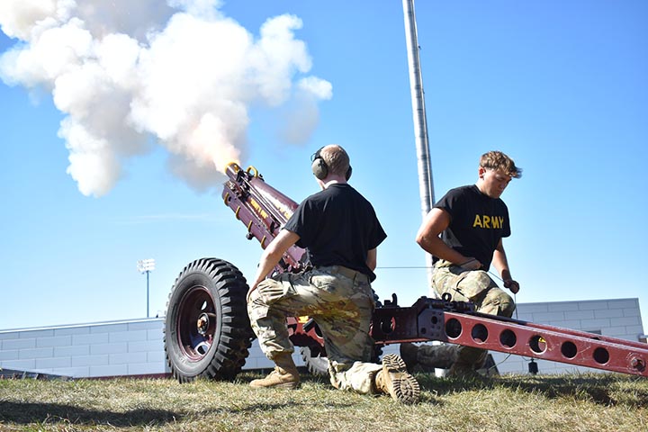 Two young men in T-shirts and camouflage pants fire a cannon; a plume of smoke is coming from the barrel.