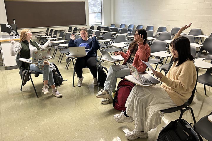 Four women collaborate on a school project while seated at desks in a classroom.