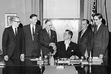 A black and white photo from 1959 shows six men standing around a table as Michigan's Governor Gerhard Williams signs a paper declaring CMU as a university.