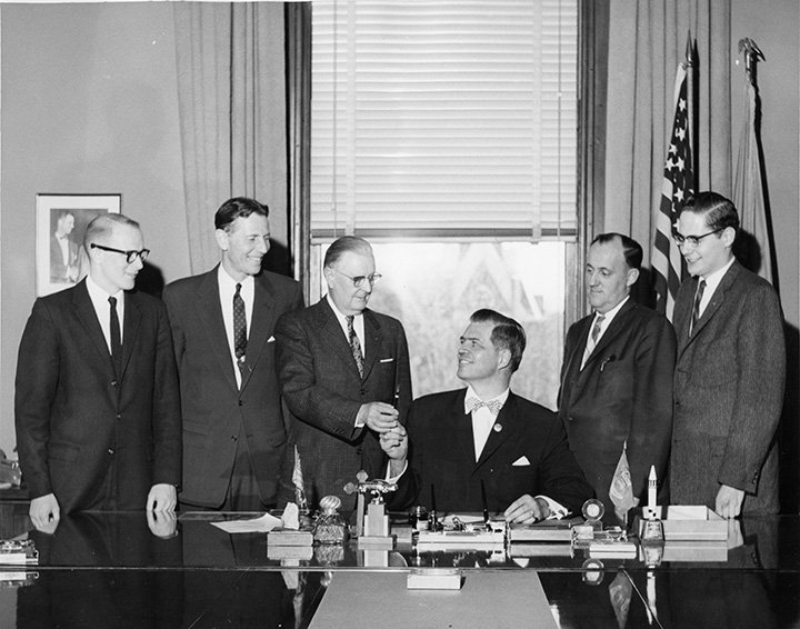 A black and white photo from 1959 shows six men standing around a table as Michigan's Governor Gerhard Williams signs a paper declaring CMU as a university.