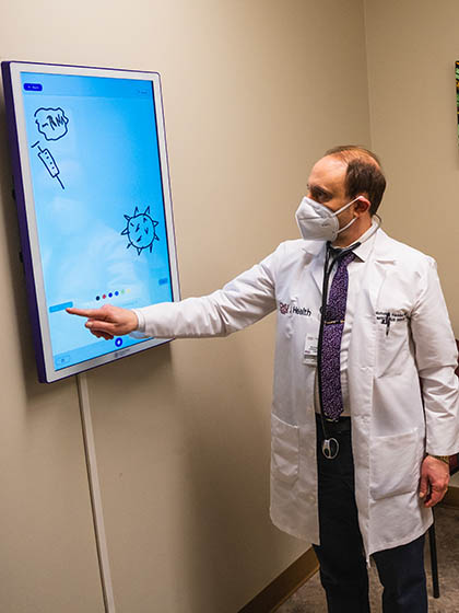 A man wearing a white coat and a light blue mask points to a section of a poster.