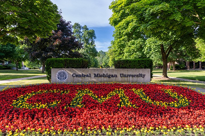 A flowerbed filled with maroon and gold flowers spells the letters C-M-U. Behind it, a cement sign with the words Central Michigan University with the university’s seal.