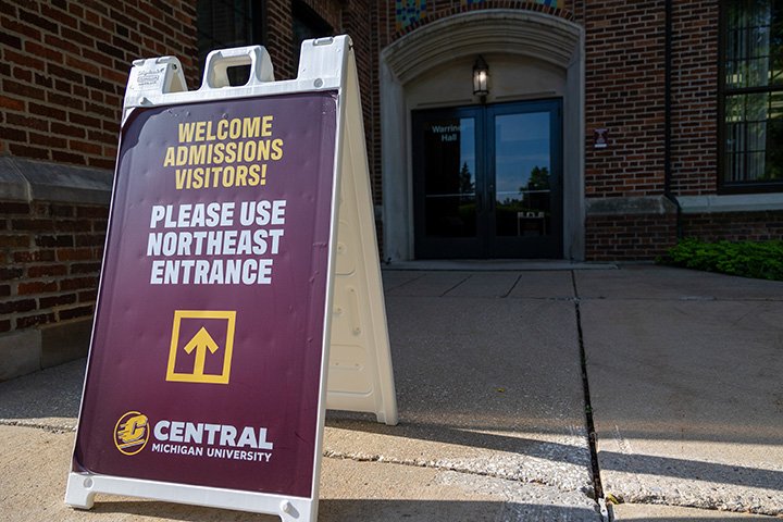 A white stand-up sign sits outside of Warriner Hall. The words Welcome Admissions Visitors, Please Use Northeast Entrance are written on the sign, pointing towards a door.