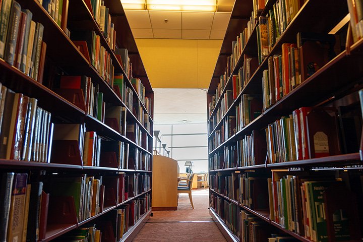 A wide-angle view in between two long bookshelves. At the center, in the distance, desk and chairs sit in front of a bright window.