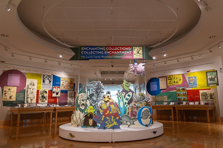 Large cutouts of children’s book characters sit on a round, circular display in the middle of a room. Children’s books in glass display cases surround the walls.