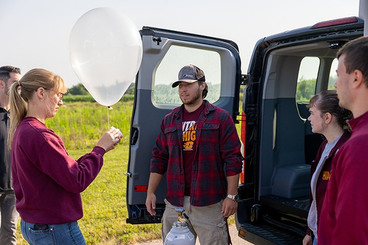 A student holds a small white balloon near the back of a van while three other students stand nearby.