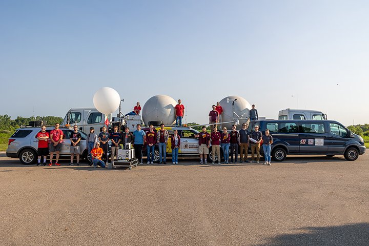 A large group of nearly 40 people pose for a group photo in a parking lot near various weather tracking vehicles.